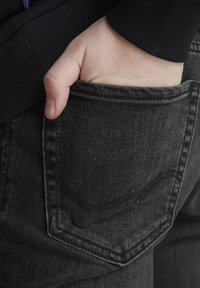 Vaqueros de mezclilla gris oscuro con un primer plano de un bolsillo trasero, mostrando una mano descansando dentro. La tela tiene una apariencia ligeramente texturizada.