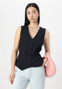 Black sleeveless vest with a V-neckline, six front buttons, and two side pockets, paired with light blue jeans and a pink handbag.