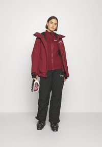 Burgunderroter Skijacke über einem passenden Oberteil, kombiniert mit schwarzen Skihosen. Schwarze Skischuhe mit rosa Akzenten. Hält Skibrille.