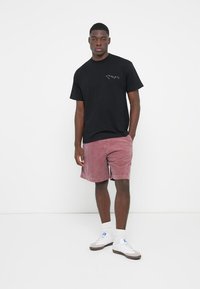 Svart kortermet t-skjorte i bomull med en liten trykket logo på brystet, kombinert med rosa kordfløyelsbukser og hvite sneakers.
