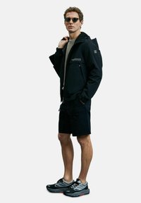 Zwarte waterdichte jas met ritssluiting aan de voorkant, capuchon en logo-details, gecombineerd met zwarte shorts en grijze sportschoenen. Accessoires zijn onder andere een zonnebril.