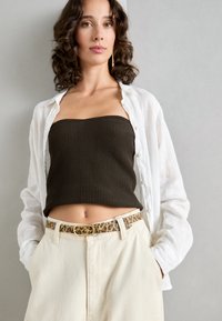 Haut sans bras côtelé marron sous une chemise en lin blanche, associé à un pantalon ivoire et une ceinture imprimé léopard avec une boucle en or.