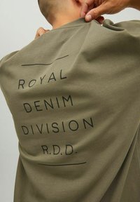 T-shirt in cotone verde oliva con grande testo nero sul retro: "ROYAL DENIM DIVISION R.D.D.", caratterizzato da un design minimalista e da una texture morbida.