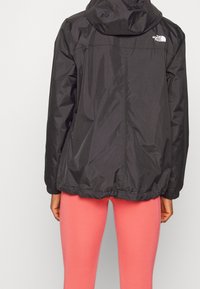 Black hooded jacket with a lightweight, smooth texture. Features elastic hem and The North Face logo on the back. Paired with pink leggings.