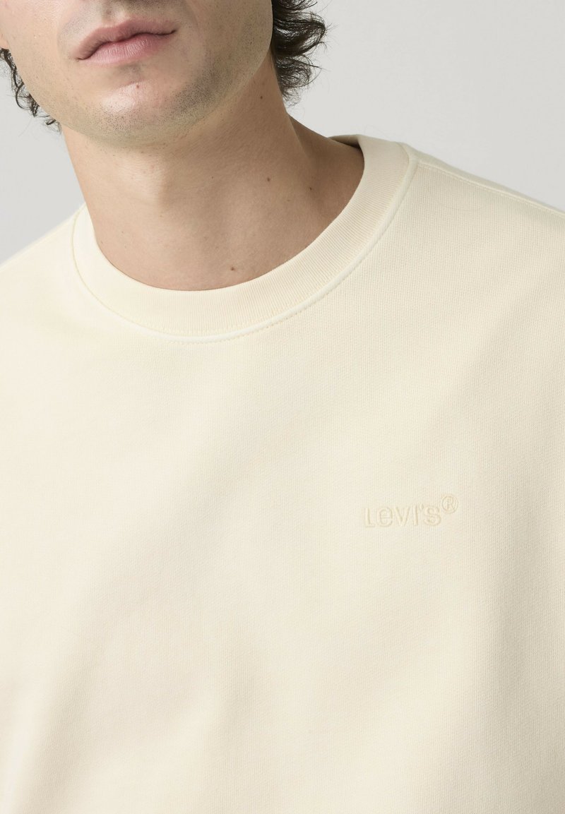Primo piano di una persona che indossa una felpa girocollo color crema con il logo Levi's discretamente ricamato sul petto a sinistra.