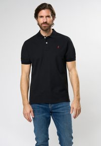 Black polo shirt with short sleeves, two-button placket, and a small red logo on the chest, paired with blue jeans. Smooth fabric texture.