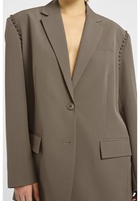 Veste blazer kaki oversize en tissu lisse, avec un col en V profond, deux poches avant et des accents cloutés le long des épaules.