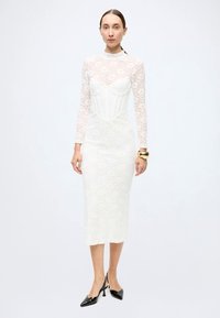 Long-sleeve white lace dress with a fitted silhouette, featuring a sheer bodice and a midi length. Black pointed toe heels and a gold bracelet accent.
