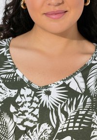 Zelený květinový top s bílým tropickým vzorem. Obsahuje výstřih do V a skladování na límci. Hladká textilie.