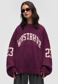 Übergroßer lila Pullover mit weißem Text "WRSTBHVA" und "23" an den Ärmeln, geripptem Muster und ausgefranstem Saum.
