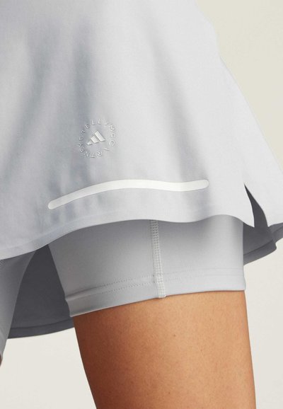 Falda deportiva gris claro, con un logotipo y una tira reflectante; superpuesta a pantalones cortos ajustados a juego con textura suave y costuras.
