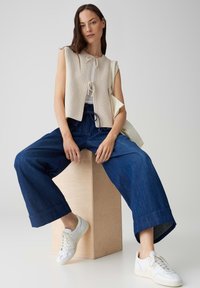 Beige gestricktes ärmelloses Top mit einer Bindeschleife, kombiniert mit blauen weit geschnittenen Jeanshosen und weißen Sneakern auf einem Holzschemel.