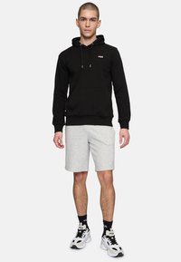 Felpa nera con cappuccio e un piccolo logo rosso, abbinata a pantaloni corti grigi e sneakers nero e bianco. Il modello è in piedi su uno sfondo neutro.