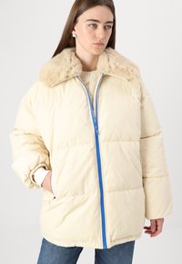 Padded cream jacket with a faux fur collar, blue zipper, and ribbed cuffs. Model wears it with blue jeans and a beige top.