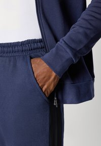 Sweatshirt desportivo azul-marinho com fecho éclair, apresentando um tecido texturizado, punhos em canelado e uma cintura elástica. Bolso incluídos nas laterais.
