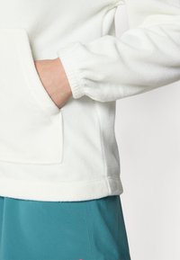 Witte fleece pullover met een kangoeroezak en ribgebreide manchetten, gecombineerd met een teal kleurige onderkant. Zachte textuur en details van een relaxte pasvorm.