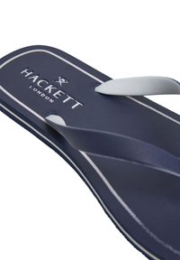 Marine flip-flop med hvide stropper, der har en struktureret sål og 'Hackett London' logo. Pyntet med hvidt kantbånd langs kanten.