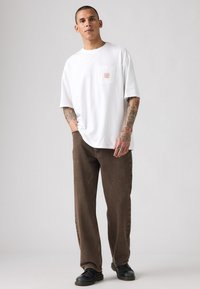 Camiseta blanca de algodón con un pequeño bolsillo de parche y un gráfico rojo, combinada con jeans marrones holgados y zapatos negros. Estilo casual, corte relajado.