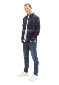 Granatowa koszula flanelowa w kratę z białymi guzikami i kieszonką na piersi, nałożona na białą koszulkę, w połączeniu z niebieskimi dżinsami i białymi trampkami.