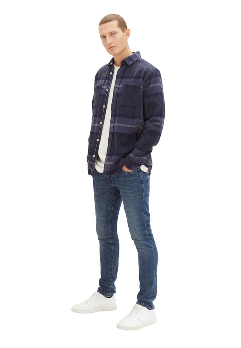 Granatowa koszula flanelowa w kratę z białymi guzikami i kieszonką na piersi, nałożona na białą koszulkę, w połączeniu z niebieskimi dżinsami i białymi trampkami.