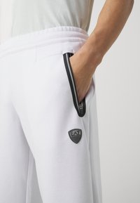 White joggers featuring a drawstring waist, zippered side pockets with black trim, and a black EA7 logo on the lower leg. Soft fabric texture.