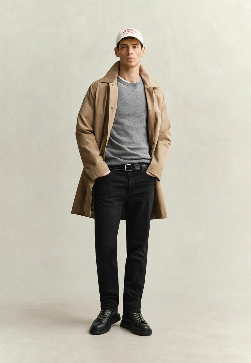 Homme portant un trench-coat beige sur un pull gris, un pantalon noir, des chaussures noires et une casquette de baseball blanche, debout les mains dans les poches.