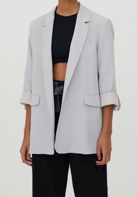 Personne portant un blazer gris clair avec manches retroussées sur un crop top noir et un pantalon noir taille haute avec une ceinture large.
