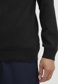 Black knitted sweater with ribbed cuffs and hem, featuring a side tag. Pairing with navy pants, showing smooth texture and fitted design.
