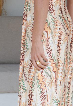 Hand met witte nagellak en gouden ring rust op oranje, groene en witte stof met bladprint van een vloeiende jurk.