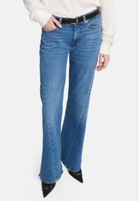 Jeans flare di denim blu chiaro con vita media, tasche frontali e una cintura nera scura. Presentano una texture liscia e cuciture visibili.