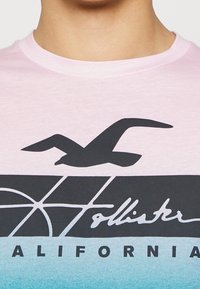 Camiseta de manga corta en degradado rosa y azul, con una silueta negra de gaviota y el texto "Hollister California" en blanco.