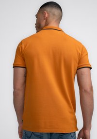Oranžové polo tričko s černým lemem kolem límce a rukávů, krátké rukávy a volný střih. Hladká bavlněná textura.