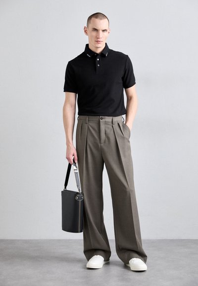 Black polo shirt, gray pleated trousers, white sneakers, holding a black cylindrical bag with a strap, against a plain gray backdrop.