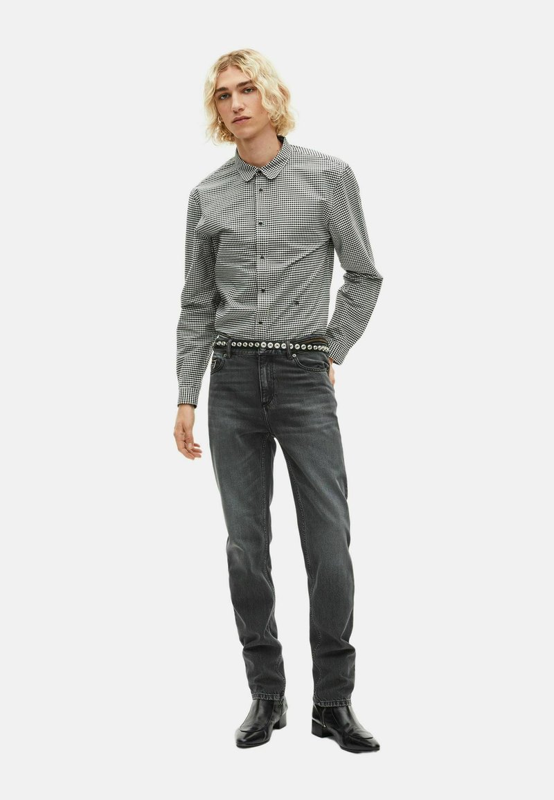 Schwarz-weiße karierte Hemd mit Button-Down-Kragen, schlank geschnitten, kombiniert mit dunkelgrauen Jeans und einem Nieten-Gürtel. Das Model trägt schwarze Knöchelstiefel.