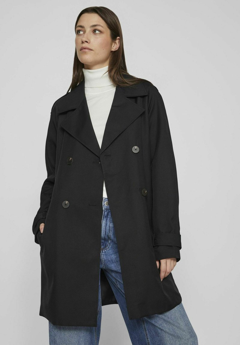 Juodas dvigubas tranšėja su įpjovimu, su dideliais mygtukais ir šoninėmis kišenėmis, dėvimas ant baltos megztinio su aukštu kaklu ir mėlynų džinsų.