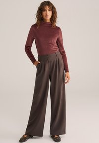 Moteris stovi vilkėdama ilgomis rankovėmis maroon spalvos marškinėlius, plačius rudus kelnes su kišenėmis ir juodas studuotas basutes ant vienodo fono.