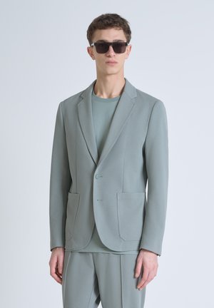 Blazer grigio chiaro realizzato in tessuto strutturato, con due tasche frontali, chiusura a un bottone e rever affilati, abbinato a una camicia coordinata.