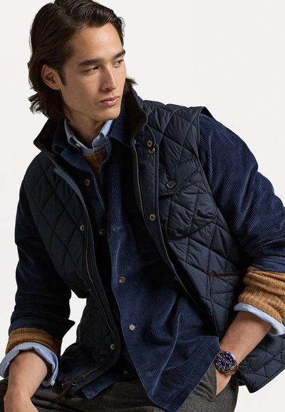 Mørkeblå quiltet vest over en blåt corduroy skjorte, lagdelt med en tan sweater. Uret har en blå urskive med metal detaljer.