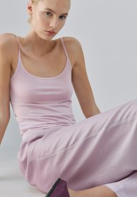 Mladá žena sediaca, oblečená v svetloružovom tielku bez rukávov a zodpovedajúcich širokých nohaviciach, s neutrálnym výrazom a minimálnym pozadím.