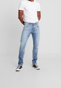 Ljusblå denimjeans med slim fit, femfickorsdesign och kontrasterande sömmar, i kombination med en enkel vit t-shirt och svarta sneakers.