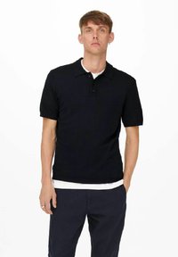 Zwarte polo met korte mouwen en een knoopsluiting met twee knopen, gedragen over een wit T-shirt met ronde hals, gecombineerd met een marineblauwe broek.