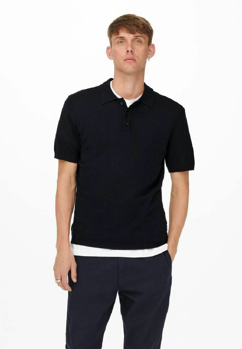 Zwarte polo met korte mouwen en een knoopsluiting met twee knopen, gedragen over een wit T-shirt met ronde hals, gecombineerd met een marineblauwe broek.