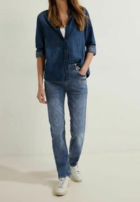 Camisa de mezclilla azul con botones y mangas enrolladas; combinada con jeans de un azul claro y zapatillas blancas. Estilo casual, con un corte relajado. Fondo liso.