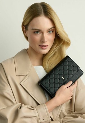 Blondynka w beżowym płaszczu trzymająca czarną pikowaną kopertówkę z małym metalowym logo z przodu na neutralnym tle.