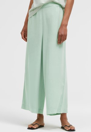 Person wearing loose light green wide-leg pants and black slide sandals standing against a plain background.