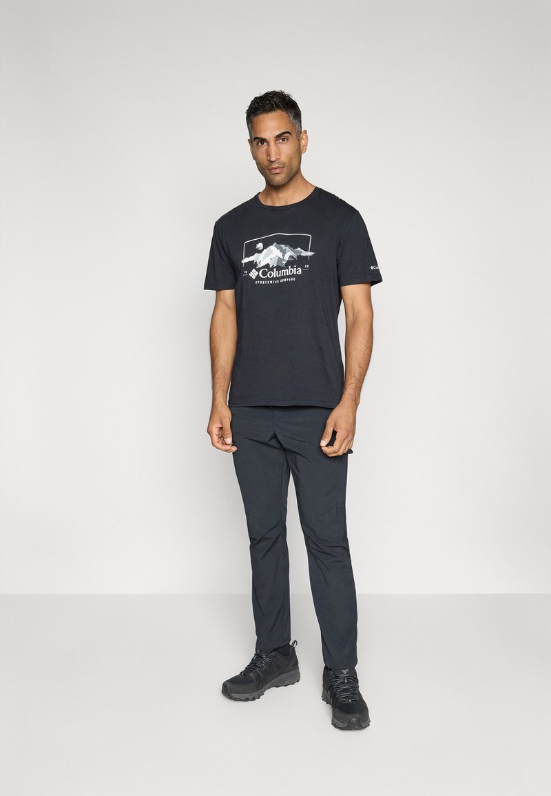 Zwarte katoenen t-shirt met een bergafbeelding in het wit, gecombineerd met marineblauwe broek en zwarte wandelschoenen, staande tegen een neutrale achtergrond.