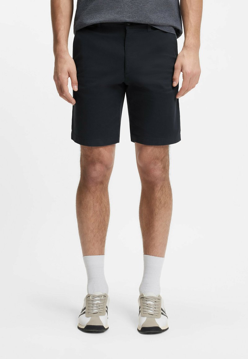 Mężczyzna w czarnych szortach, białych krótkich skarpetach i białych sportowych butach z szaro-czarnymi detalami, stojący na jednolitym białym tle.