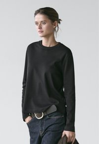 Ung kvinde iført en sort langærmet skjorte og mørke jeans med hænderne i lommen, stående mod en ensfarvet lys baggrund.