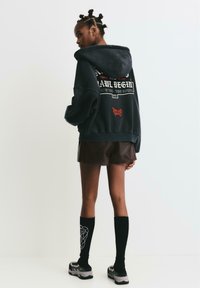 Tmavo šedá mikina s grafikou a červenými akcentmi, kombinovaná s hnedou sukňou, čiernymi podkolienkami a robustnými teniskami.