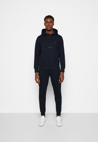 Felpa blu navy con cappuccio e lacci, tasca a marsupio e polsini a coste, abbinata a pantaloni jogger coordinati e sneakers bianche.